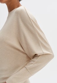 Haut beige texturé à coupe décontractée, avec un col montant et des épaules tombantes. Motif côtelé, tissu doux, manches longues.