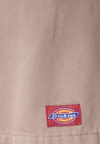 Tessuto beige con trama diagonale; presenta un'etichetta rossa con il logo "Dickies" in bianco e blu ricamato.
