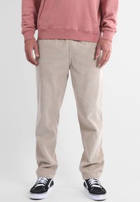Pantalon en velours côtelé beige à côtes verticales, coupe décontractée, avec poches latérales, associé à des baskets noires et un sweat-shirt rose.