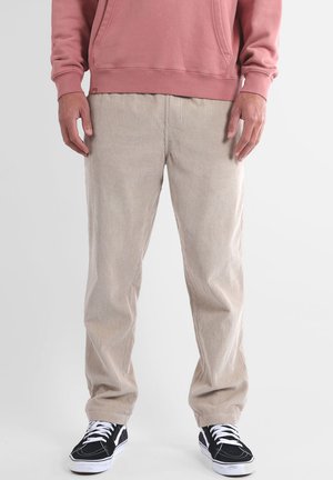 Pantalon en velours côtelé beige à côtes verticales, coupe décontractée, avec poches latérales, associé à des baskets noires et un sweat-shirt rose.