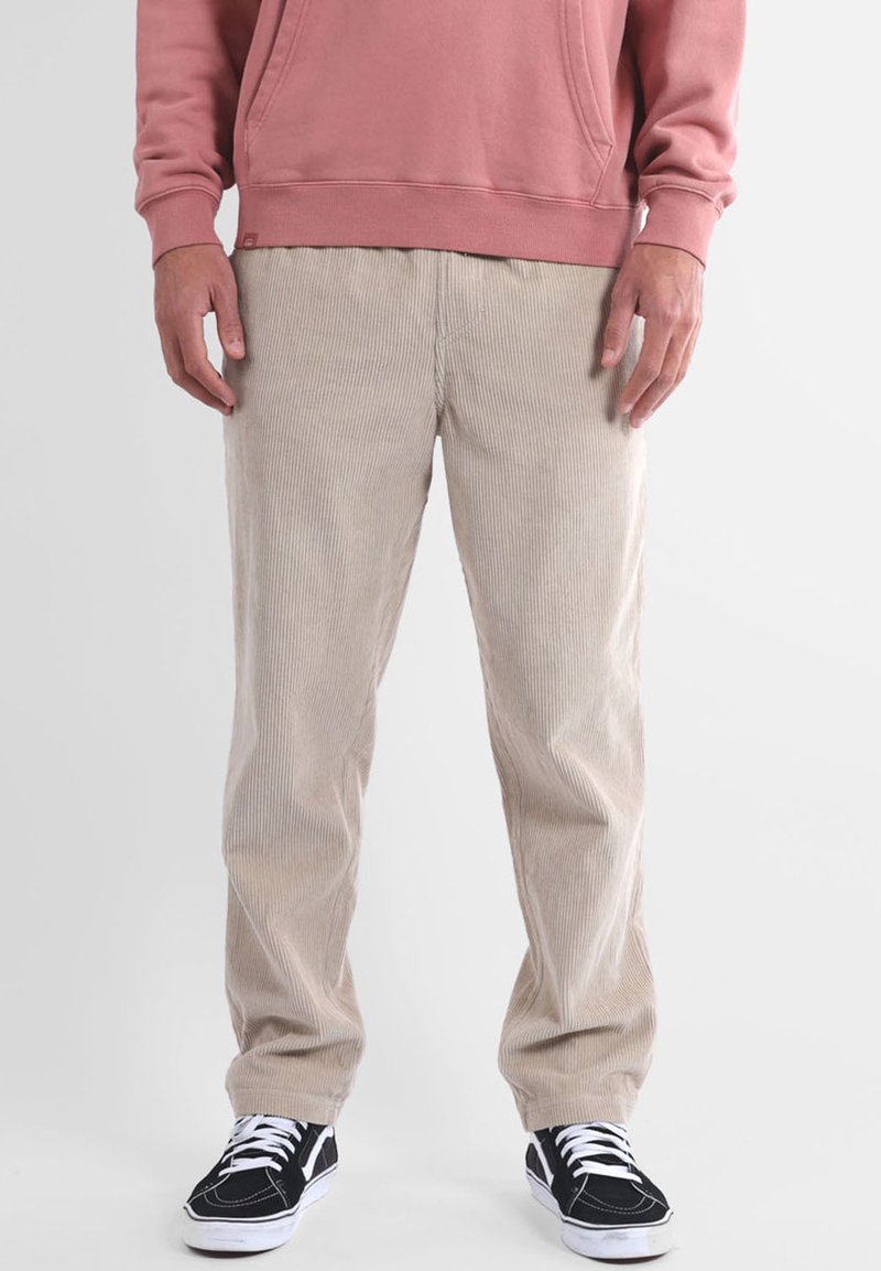 Pantalon en velours côtelé beige à côtes verticales, coupe décontractée, avec poches latérales, associé à des baskets noires et un sweat-shirt rose.