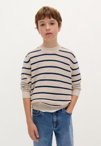 Beiger Strickpullover mit marineblauen Streifen, Rundhalsausschnitt und gerippten Bündchen. Kombiniert mit blauen Jeans. Das Modell steht mit den Händen in den Taschen.