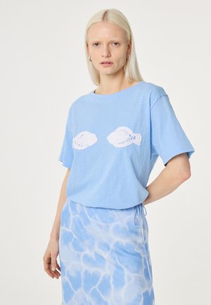 Blonde Frau trägt ein hellblaues T-Shirt mit weißen Fischmotiven, kombiniert mit einem blau-weißen Rock mit Wasser-Muster, steht vor einem weißen Hintergrund.