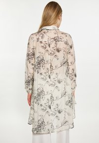 Blusa lunga e trasparente con stampa floreale in crema e disegni di fiori neri; presenta un colletto, polsini abbottonati e un orlo leggermente curvato.