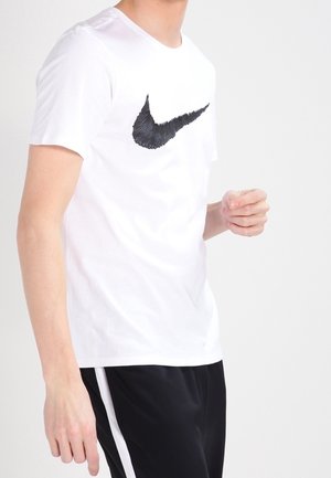 Młoda osoba w białej koszulce z dużym czarnym logo Nike oraz czarnych spodniach sportowych z białym paskiem, na jednolitym tle.