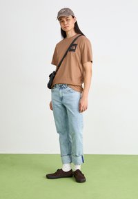Bruine t-shirt met korte mouwen en logo op de borst, lichtblauwe jeans, zwarte schoudertas en bruin schoenen met witte sokken. Eenvoudig ontwerp, casual look.