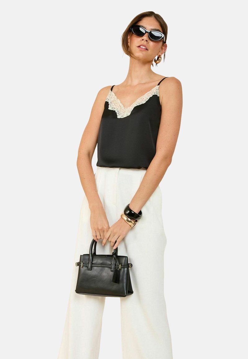 Camisole in seta nera con rifiniture in pizzo, abbinato a pantaloni larghi bianchi. La modella tiene una piccola borsa a tracolla in pelle nera, indossando occhiali da sole neri.