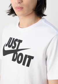 Hvid bomulds T-shirt med et iøjnefaldende sort "JUST DO IT" slogan og en Nike swoosh. Kortærmet, rund halsudskæring, glat tekstur.