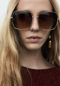 Oversized brown gradient sunglasses with a gold metal frame, featuring square lenses and thin temples, worn with loose, wavy blonde hair.