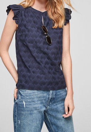 Kvinde iført en mørkeblå, flæset ærmeløs top med tekstureret mønster og solbriller hængende fra udskæringen, kombineret med lyseblå jeans.