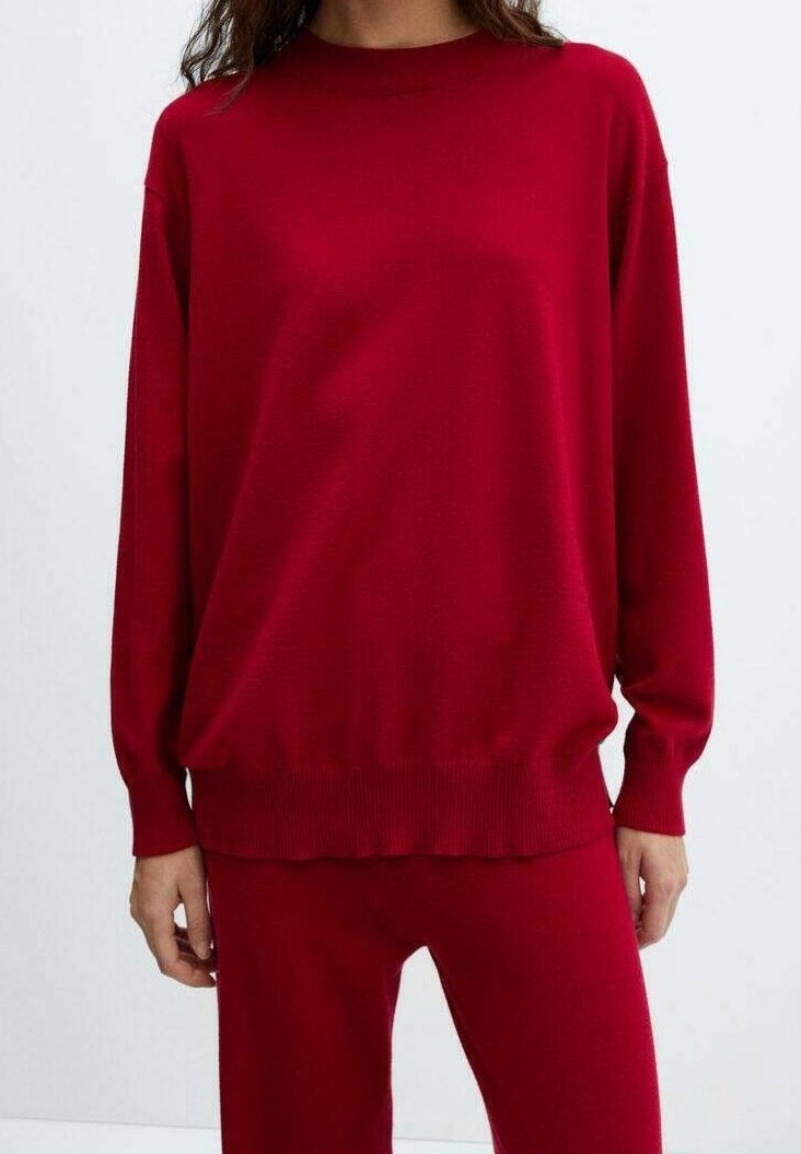 Femme portant un pull rouge uni à manches longues et un pantalon rouge assorti, debout devant un fond uni clair.