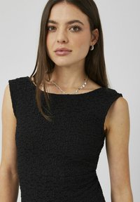 Robe noire en tissu texturé, avec un col bateau et des manches courtes. Un collier argenté orné de pendentifs circulaires complète le look.