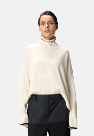 Suéter de punto color crema con cuello alto y textura acanalada, con hombros caídos, combinado con pantalones negros.