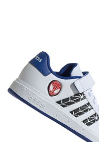 Baskets blanches avec des accents bleus et un design en motif de toile noir. Présente un logo Spider-Man sur le côté et une sangle en Velcro. Semelle en caoutchouc.