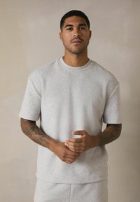 Helles graues T-Shirt mit kurzen Ärmeln aus strukturiertem Stoff, mit Rundhalsausschnitt und lockerer Passform. Sichtbare Tätowierungen an den Armen.