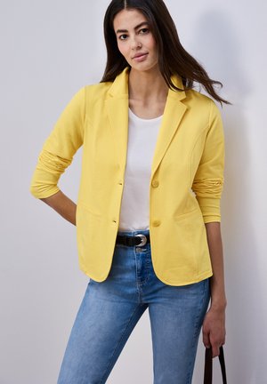 Mujer con blazer amarillo, camisa blanca y vaqueros azules, de pie con una mano en el bolsillo y sosteniendo una correa negra con la otra.