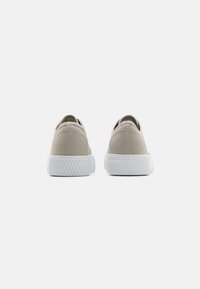 Des baskets en toile beige avec un dessus texturé, une semelle en caoutchouc blanche et un design bas. Elles présentent un bout arrondi et des accents minimalistes.