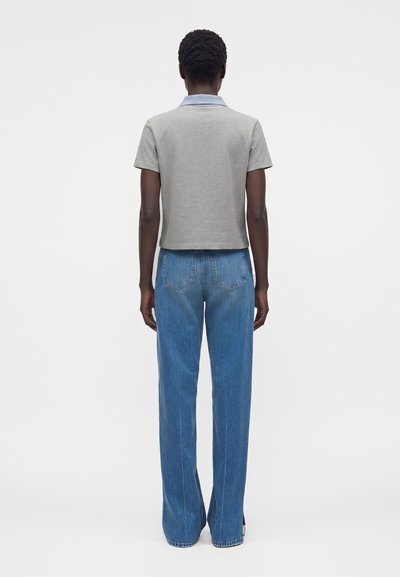 Άτομο που στέκεται με την πλάτη γυρισμένη, φορά ένα ανοιχτό γκρι κοντομάνικο πουκάμισο με γιακά και μπλε τζιν παντελόνι με ίσια μπατζάκια.