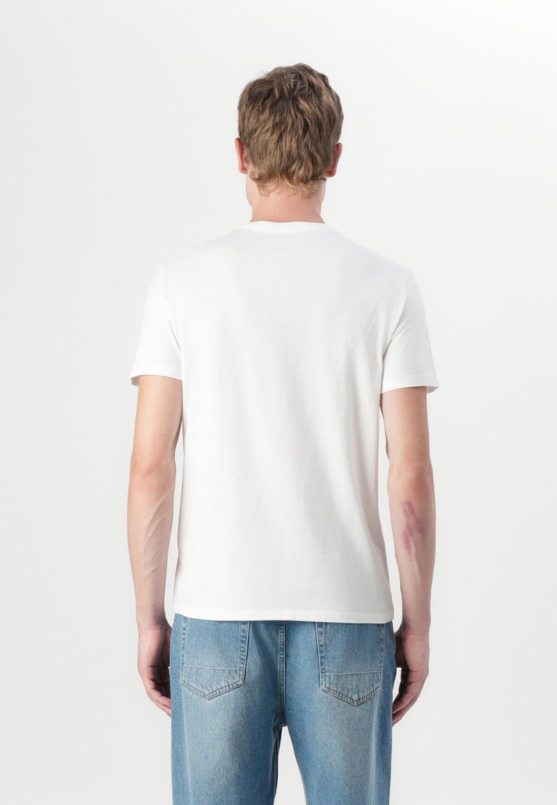 Camiseta de algodón blanca con escote redondo, mangas cortas y corte relajado; llevada con jeans azules deslavados, mostrando vista trasera.