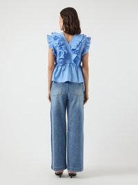 Kvinne iført en blå bluse med volanger uten ermer og V-hæl, kombinert med brede blå jeans og svarte hælede sandaler, vendt bort.