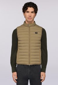 Hnědá prošívaná vesta s horizontálním prošíváním, vysokým límcem a předním zipem. Nositá přes tmavě zelenou košili s dlouhým rukávem, která má logo jako akcent.