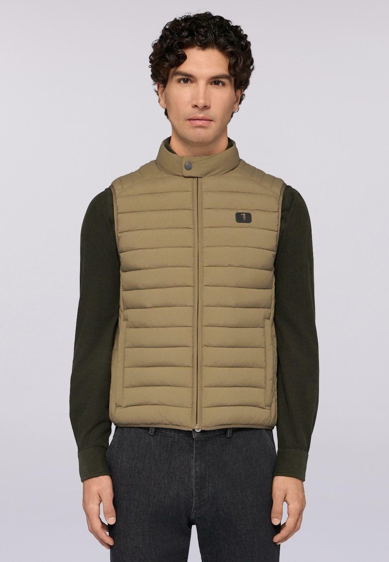 Hnědá prošívaná vesta s horizontálním prošíváním, vysokým límcem a předním zipem. Nositá přes tmavě zelenou košili s dlouhým rukávem, která má logo jako akcent.