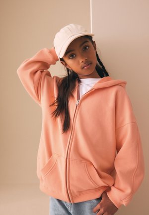 Young girl with long dark hair in braids wears a light beige cap, peach zip-up hoodie, and light blue jeans, posing with one hand behind her head.