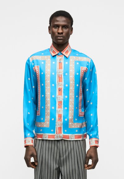 Muž nosiaci žiarivo modrú a oranžovú vzorovanú košeľu na gombíky s geometrickými a kvetinovými motívmi, doplnenú o čierne a biele nohavice s vertikálnymi pruhmi.