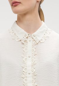 Blouse blanche avec un col en dentelle, boutonnage à l'avant et tissu texturé. Détail du motif en dentelle autour de l'encolure bien mis en valeur.