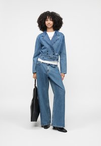 Chaqueta de mezclilla con diseño de doble botonadura, cintura ceñida por un cinturón y jeans de pierna ancha. Ambos son de un azul claro, combinados con una camisa blanca y un bolso negro.