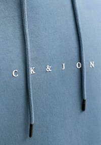 Hele sinine kapuutsiga dressipluus pehme tekstuuriga, esiküljel valgete tähtedega kujundus "CK & JON" ning mustade otstega punutud nöörid.