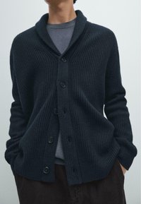Dark ribbed knit cardigan with a shawl collar, seven large buttons, and a relaxed fit, worn over a gray crew-neck shirt.