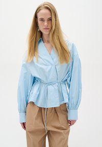 Light blue button-up shirt with a drawstring waist, balloon sleeves, and a glossy texture, paired with loose tan trousers.
