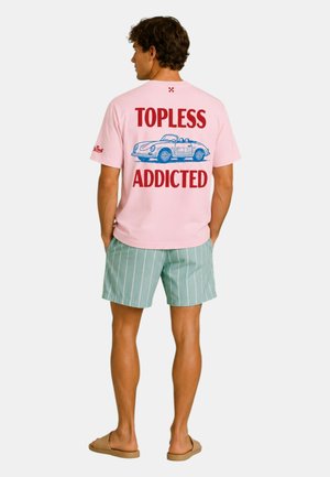 Uomo con maglietta rosa con testo "TOPLESS ADDICTED" e grafica di auto decappottabile blu, pantaloncini a righe verde menta e sandali beige.