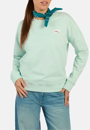 Vrouw draagt een lichtgroene Reebok-trui, een blauwe sjaal met patroon om haar nek en een blauwe spijkerbroek, met één hand in haar zak en de andere op haar heup.