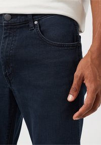 Närbild av en person som bär mörkblåjeans med en synlig framficka, knapp och en avslappnad hand vid låret.