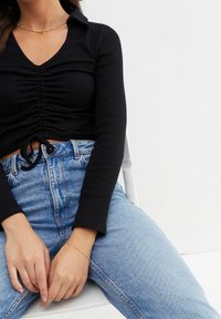 Schwarzes, geripptes Langarmoberteil mit V-Ausschnitt und Kordelzug in der Taille, kombiniert mit hellblauen, hoch taillierten Jeans. Sichtbares goldfarbenes Armband.