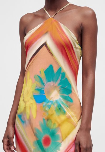 Γυναίκα που φοράει πολύχρωμο φόρεμα με halter neck και μεγάλα αφηρημένα μπλε, κίτρινα και κόκκινα λουλουδένια σχέδια σε απαλό πορτοκαλί φόντο.