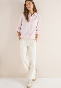 Kobieta stojąca przy ścianie, ubrana w jasnoróżową koszulę zapinaną na guziki, białe szerokie spodnie i białe trampki z niebieskimi akcentami.