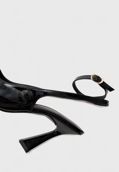 Black patent leather high-heeled sandals with an open toe, angular heel, and thin ankle strap featuring a circular gold buckle detail.