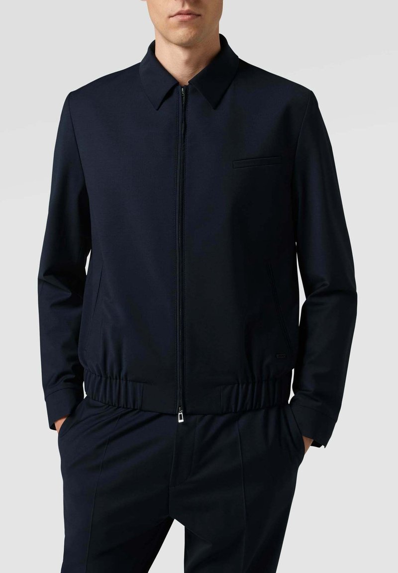 Giacca bomber blu navy con zip frontale, colletto a punta, orlo elasticizzato e taschino sul petto; realizzata in tessuto morbido con finitura opaca.