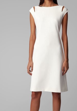 Frau trägt ein ärmelloses weißes knielanges Kleid mit Ausschnitten an den Schultern vor einem schlichten grauen Hintergrund.