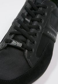 Czarny sneaker z mieszanką teksturowanej skóry i zamszu, z okrągłymi czarnymi sznurówkami, markową metką i gładką gumową podeszwą.