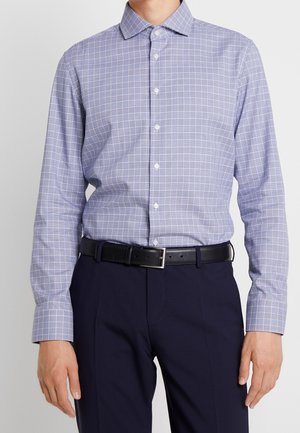 Mężczyzna w jasnofioletowej kraciastej koszuli z długim rękawem, wsadzonej w ciemnogranatowe spodnie z czarnym skórzanym paskiem i srebrną klamrą.