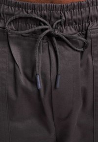 Pantalones marrón oscuro con cinturilla elástica y cordón. La tela es suave y tiene un brillo sutil, con bolsillos laterales y detalles cosidos.