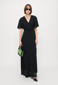 Black maxi dress with short, flowing sleeves, wrap front, and a smooth texture. The model holds a green beaded handbag and wears black shoes.