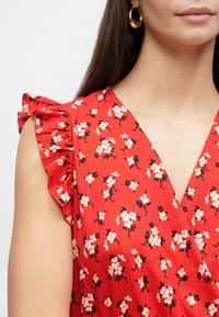 Rochie florală roșie din material texturat, cu mâneci cu volane și decolteu în V, decorată cu modele mici de flori albe și negre.