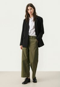 Zwarte blazer, witte shirt en olijfgroene wijde broek met een gestructureerde afwerking. Zwarte veterschoenen maken de look compleet.
