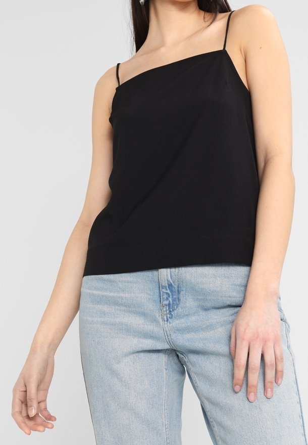 Femme portant un débardeur noir à une épaule avec des fines bretelles et un jean taille haute bleu clair sur un fond uni.