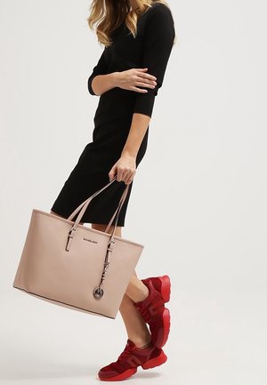 Femme en robe noire tenant un grand sac cabas beige, portant des baskets rouge vif, debout devant un fond clair uni.